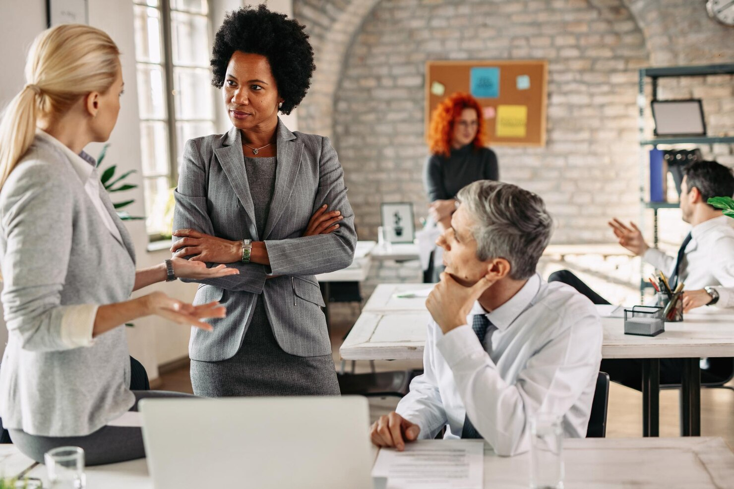 A man, seated, and two women, standing, in a business meeting in an open-concept office.